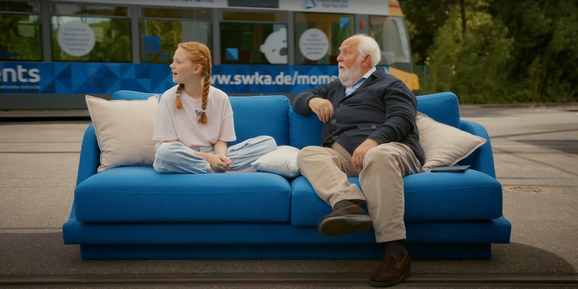 Szene aus dem Imagefilm für die Stadtwerke Karlsruhe: Ein Mädchen und ein älterer Mann sitzen auf einem blauen Sofa vor einer Straßenbahn